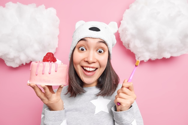 Mujer sonríe con dientes vestida con pijama sostiene un sabroso pastel y un cepillo de dientes tiene caries debido a comer demasiados dulces aislados en rosa Foto gratis