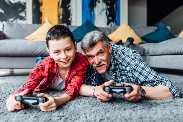 Niño sonriente y su abuelo jugando videojuegos juntos en casa acostado