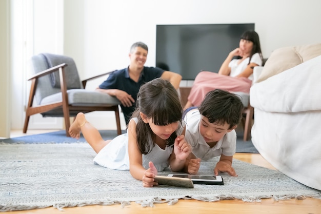 Padre feliz viendo a los niños pequeños tirados en el piso en la sala de estar y usando aparatos digitales juntos. Foto gratis