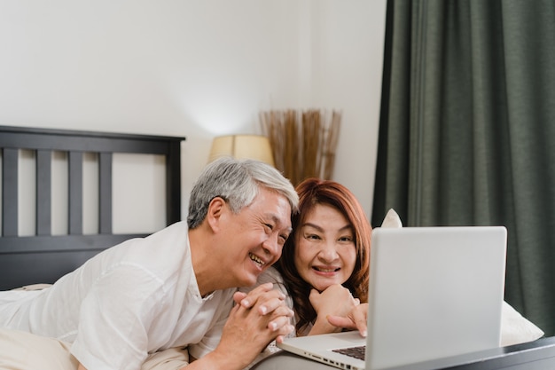 Pares mayores asiáticos que usan la computadora portátil en el país. los abuelos, marido y esposa chinos mayores asiáticos felices después de despertarse, viendo la película acostada en la cama en el dormitorio en casa en el concepto de mañana. Foto gratis