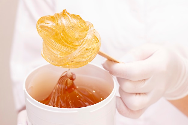 Pasta De Azucar Liquida O Cera Para La Depilacion En Un Palo De Madera De Primer Plano Sobre Un Blanco Contra Los Uniformes De Un Maestro De Depilacion Foto Premium