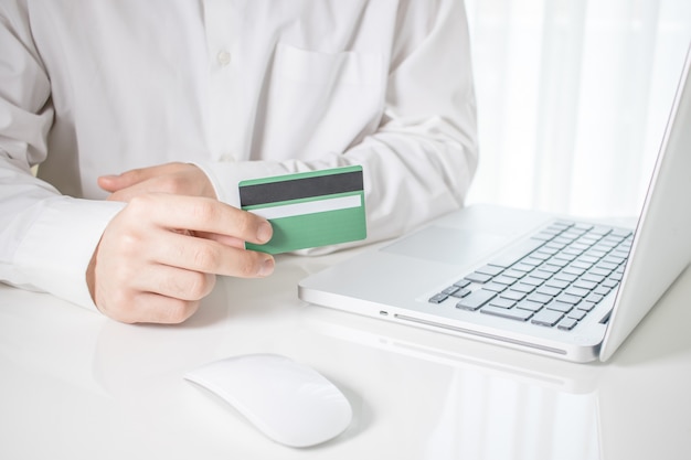 Persona que tiene una tarjeta de crédito verde con una computadora portátil y un mouse de computadora sobre una mesa blanca Foto gratis