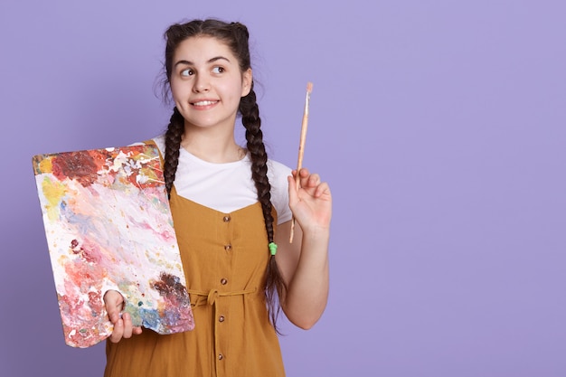 La pintora feliz joven piensa en un nuevo proyecto, tiene una expresión facial pensativa, mirando sonriendo a un lado, posando aislada sobre la pared lila. copie el espacio. Foto gratis
