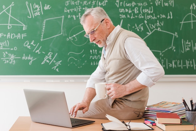 Profesor mayor que se sienta en el escritorio y que pulsa en la computadora portátil Foto gratis