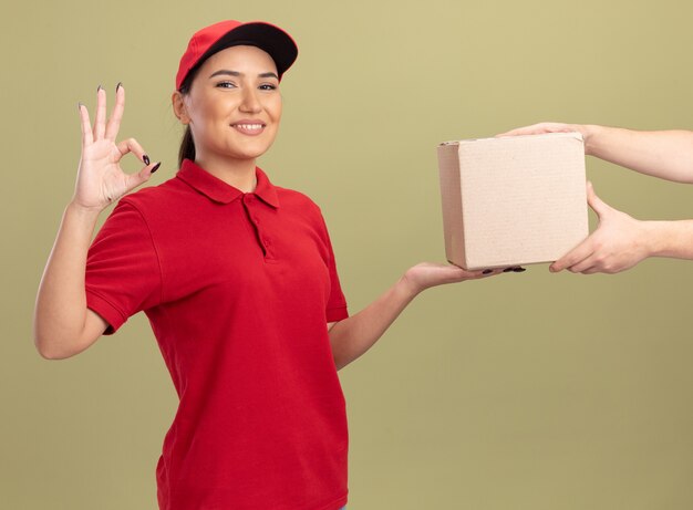 Repartidor Joven En Uniforme Rojo Y Gorra Dando Caja De Cartón A Un Cliente Sonriendo Amable 4461