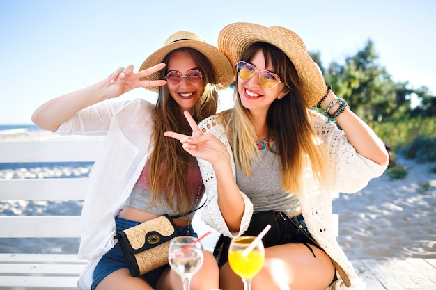 Retrato al aire libre de las chicas hipster divertidas felices de la compañía volviéndose locas en el café de la playa, bebiendo deliciosos cócteles riendo y sonriendo, trajes de verano boho brillante vintage, relaciones y diversión. Foto gratis