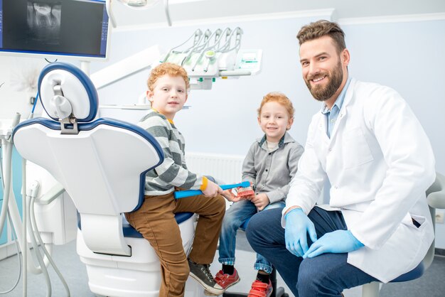 Retrato De Guapo Dentista Y Niños Felices Sentados En La Silla En El Consultorio Dental Foto