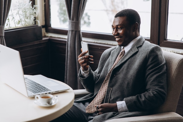 Retrato del hombre de negocios africano feliz que usa el teléfono mientras que trabaja en la computadora portátil en un restaurante. Foto Premium