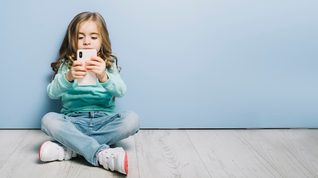 Retrato de una niÃ±a sentada en el piso de madera dura mirando smartphone Foto gratis