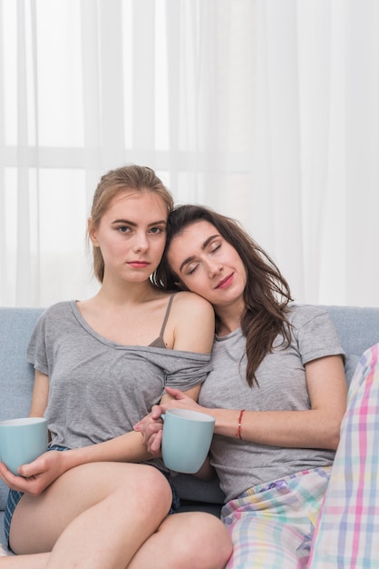 Retrato De Una Pareja De Lesbianas Jovenes Sentados En El Sofa Sosteniendo Una Taza De Cafe Azul Foto Gratis