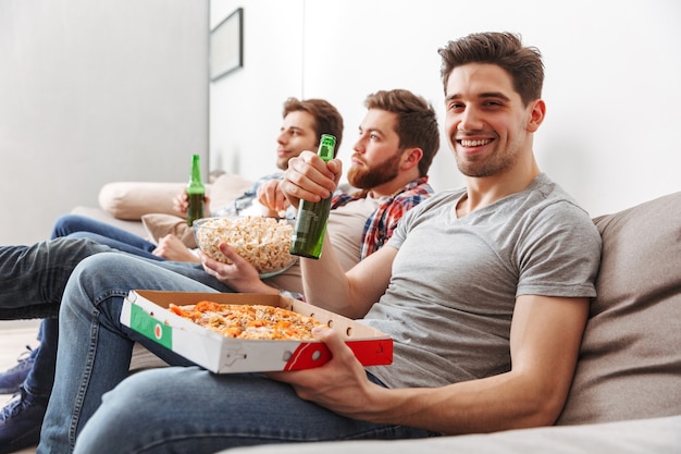 Retrato De Tres Hombres Jóvenes Felices Viendo Fútbol Mientras Está Sentado En Casa Con Cerveza 