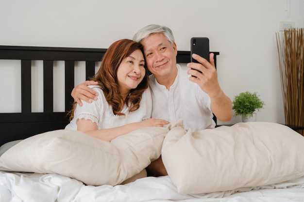 Selfie de pareja senior asiática en casa. abuelos, marido y esposa chinos mayores asiáticos felices usando el selfie del teléfono móvil después de despertarse acostado en cama en dormitorio en casa en el concepto de la mañana. Foto gratis