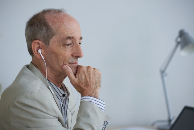 Senior empresario caucásico sentado en la oficina con auriculares y mirando portátil Foto gratis