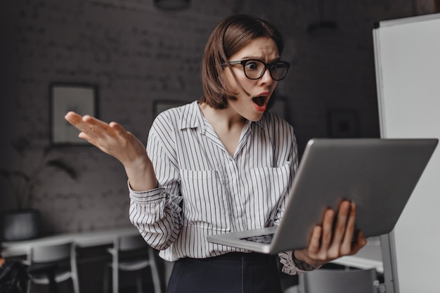 Señora de negocios feroz mira indignada y conmocionada en la computadora portátil y extiende sus brazos contra el fondo del lugar de trabajo blanco. Foto gratis