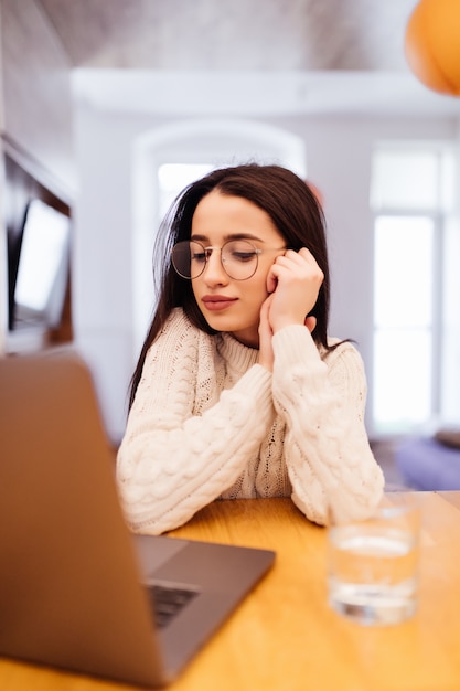 Soñadora dama de suéter blanco está sentada en la cocina y trabajando en su computadora portátil