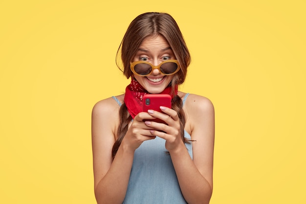 Sonriente mujer de cabello oscuro con expresión alegre, sostiene un teléfono móvil rojo, feliz de leer un mensaje de texto, conectado a internet inalámbrico, aislado sobre una pared amarilla. gente, tecnología, ocio Foto gratis