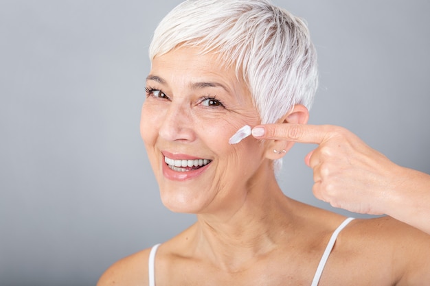 Sonriente mujer senior aplicando loción antienvejecimiento para eliminar las ojeras bajo los ojos. mujer madura con