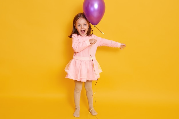Tiro horizontal de niña con globo morado aislado sobre amarillo todas las niñas gritando algo, celebrando el día de la hierba, niño vestido de rosa y con el pelo oscuro. Foto gratis