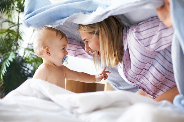 Tiro de vista lateral de la encantadora joven alegre en pijama jugando al escondite con la hija del niño. adorable niño lindo bebé chupando chupete mirando a la madre, con expresión facial juguetona Foto gratis