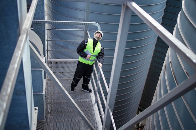 Trabajador de fábrica de pie sobre una plataforma metálica entre tanques de almacenamiento industrial y mirando hacia arriba para la inspección visual de los silos de producción de alimentos Foto gratis