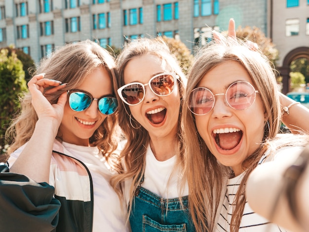 Tres jóvenes sonrientes mujeres hipster en ropa de verano. chicas tomando fotos de autorretrato en smartphone. modelos posando en la calle. mujeres mostrando emociones positivas en las gafas de sol. Foto gratis