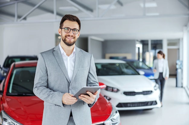 Vendedor De Coches Amistoso Sonriente En Traje De Pie En El Salón Del Coche Y Sosteniendo La 