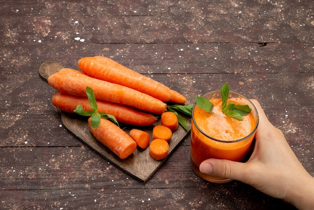 Vista frontal de jugo de zanahoria fresco dentro de un vaso largo con hojas y junto con zanahorias frescas en el escritorio marrón Foto gratis