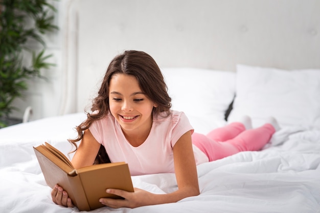Vista frontal niña sonriente en casa leyendo en la cama | Foto Gratis