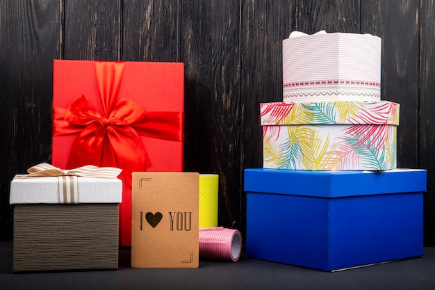 Vista lateral de una pila de cajas de regalo coloridas y una pequeña tarjeta de te amo en la mesa de madera oscura Foto gratis