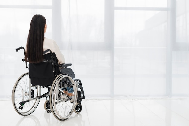 Vista posterior de una mujer sentada en silla de ruedas mirando a la ventana Foto gratis