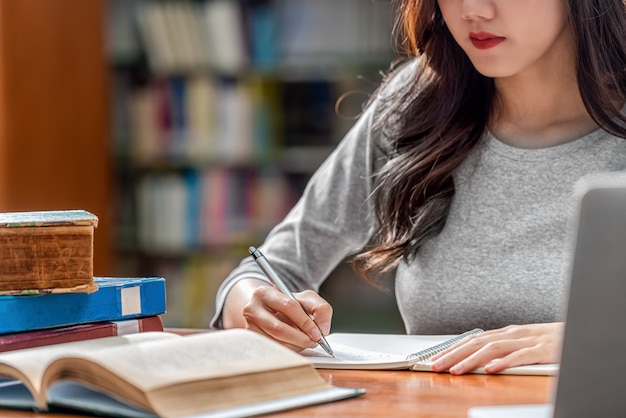 Asian giovane studente a mano a scrivere i compiti e utilizzando la tecnologia portatile nella biblioteca dell'università o colleage con vari libri e fermo sopra lo scaffale Foto Premium
