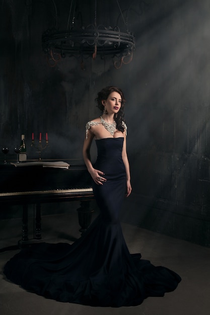 Bella giovane donna in abito nero accanto a un pianoforte con candele candelabri e vino, atmosfera drammatica scuro del castello. boemia. Foto Premium