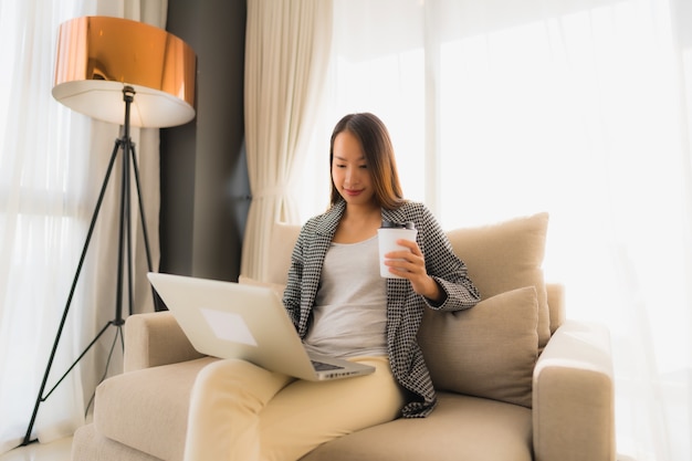 Bello giovane ritratto asiatico facendo uso del computer e del computer portatile con la tazza di caffè che si siede sul sofà Foto Gratuite