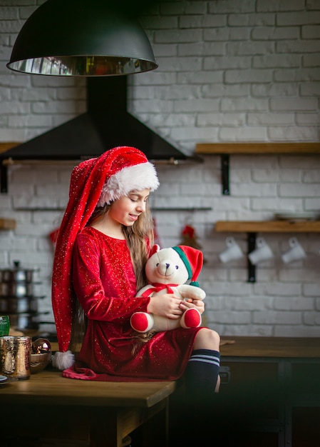 Buon Natale 105.Buon Natale La Bambina Sveglia In Cappello Di Santa Sta Tenendo Un Orsacchiotto Che Si Siede Nella Cucina Aspettando La Vacanza Foto Premium