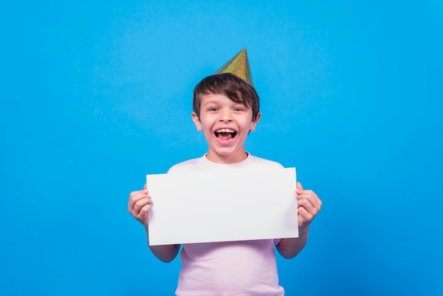Cappello d'uso del partito del ragazzino emozionante che giudica carta in bianco a disposizione sulla superficie del blu Foto Gratuite
