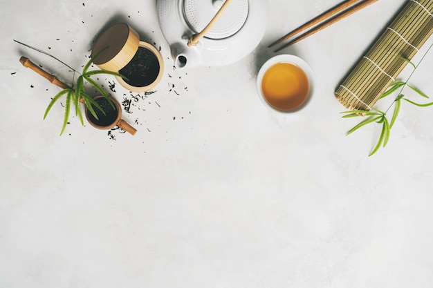 Concetto di tè asiatico, due tazze bianche di tè, teiera, set da tè, bacchette, stuoia di bambù circondato con tè verde secco su sfondo bianco Foto Premium