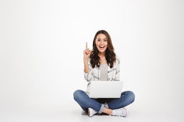 Donna felice del brunette in camicia che si siede sul pavimento con il computer portatile mentre avendo idea ed esaminando la macchina fotografica sopra grey Foto Gratuite