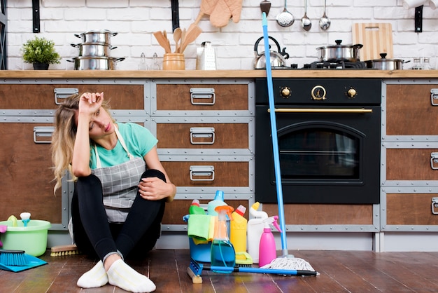 Donna stanca che si siede sul pavimento della cucina con prodotti e attrezzature per la pulizia Foto Gratuite