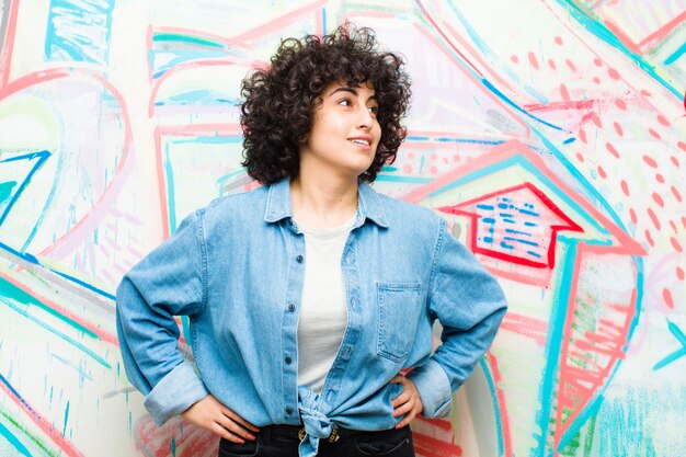 Giovane donna afro graziosa che sembra allegra felice e sicura che sorride fiero e che guarda da parte con entrambe le mani sulle anche contro la parete dei graffiti Foto Premium