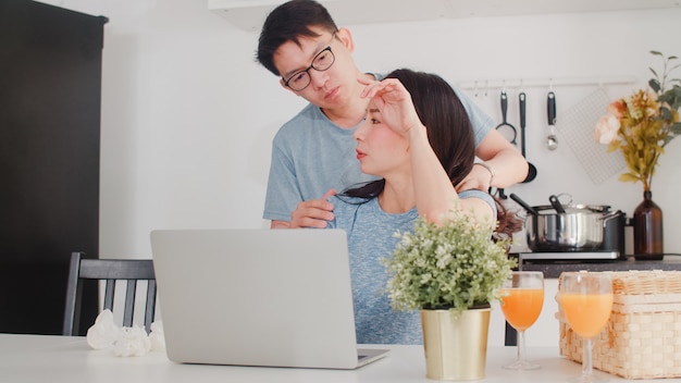 Giovane donna di affari asiatica seria, stress, stanca e malata mentre si lavora al computer portatile a casa. il marito le dà il bicchiere d'acqua mentre lavora sodo nella cucina moderna a casa la mattina. Foto Gratuite