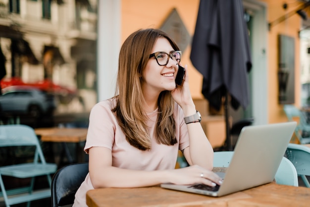 Giovane donna sorridente che parla sul telefono e che scrive sul computer portatile nel caffè all'aperto Foto Premium