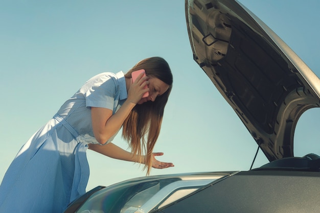 Giovane e bella ragazza vicino a un'auto rotta con un cofano aperto. problemi con la macchina, non si avvia, non funziona. Foto Premium