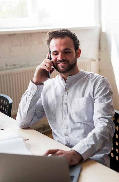 Giovane sorridente parlando sul cellulare sul posto di lavoro Foto Gratuite