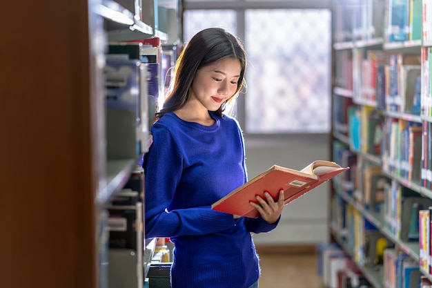 Giovane studentessa asiatica in abito casual in piedi e leggendo il libro allo scaffale del libro nella biblioteca dell'università o colleage con varie pareti del libro, torna al concetto di scuola Foto Premium