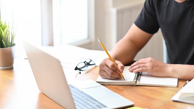 Hs dell'uomo del primo piano che scrive sul computer portatile della carta del taccuino. Foto Premium