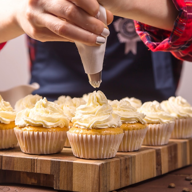 Il pasticcere femminile in grembiule blu e una camicia scozzese rossa applica la crema ai cupcakes Foto Premium