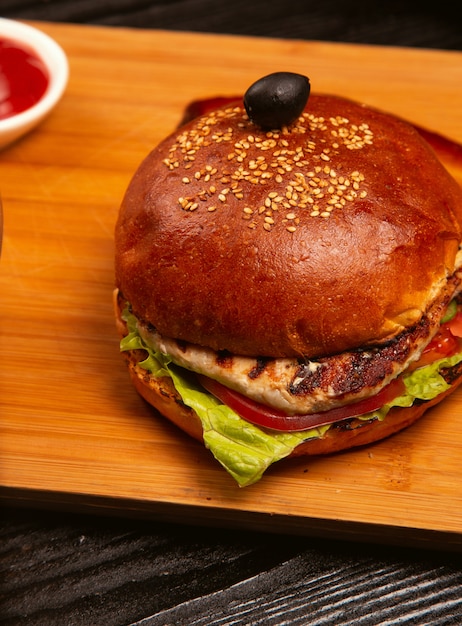 L'hamburger di carne di pollo con pomodoro e lattuga all'interno è servito con olive nere e ketchup su un vassoio di legno. Foto Gratuite