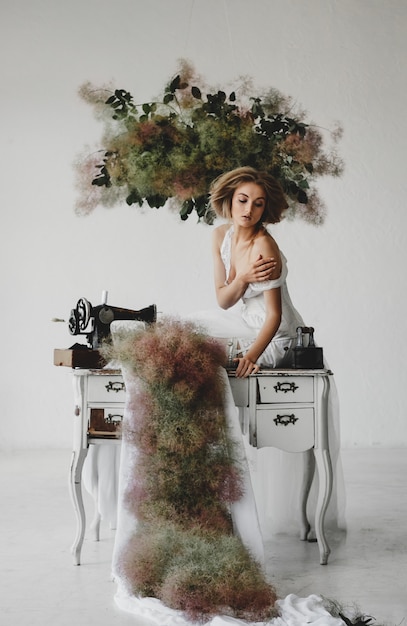 La donna in vestiti bianchi si siede in una stanza con i fiori e la macchina per cucire Foto Gratuite