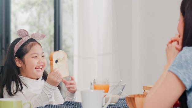 La famiglia giapponese asiatica fa colazione a casa. la figlia asiatica seleziona e gioca il sorriso di risata del pane con i genitori mentre mangia il cereale e il latte dei fiocchi di mais in ciotola sulla tavola in cucina moderna di mattina. Foto Gratuite