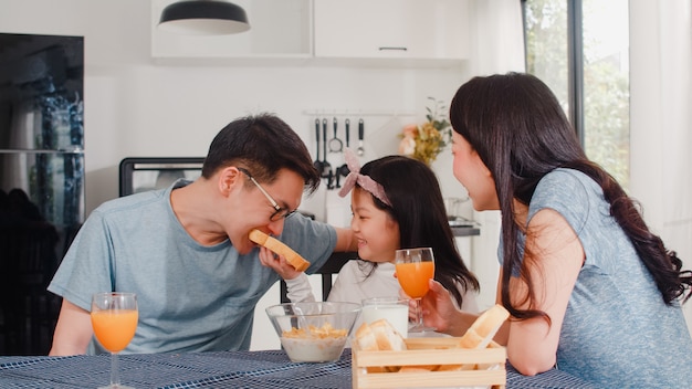 La famiglia giapponese asiatica fa colazione a casa. la mamma, il papà e la figlia asiatici che si sentono felici parlano insieme mentre mangiano il pane, i cereali dei fiocchi di mais e il latte in ciotola sulla tavola nella cucina di mattina. Foto Gratuite